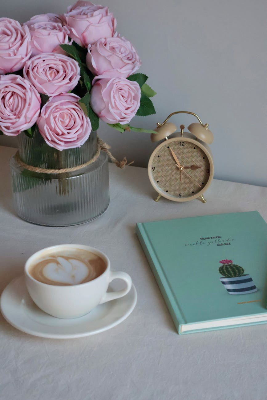 Artificial roses in vase, alarm clock, notebook and a cup of coffee on a desk